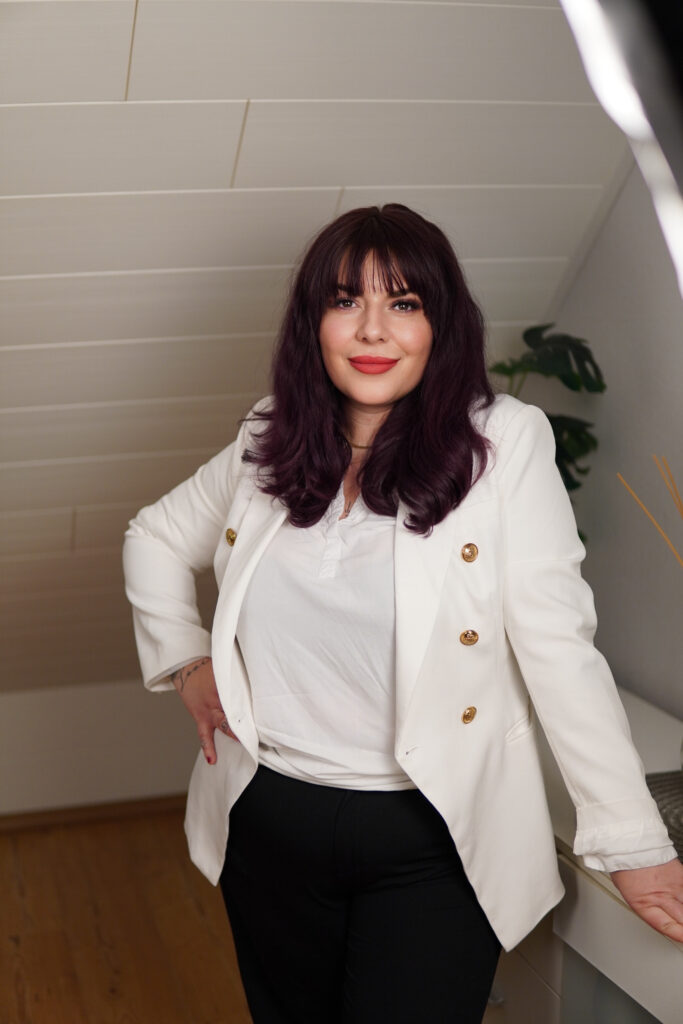 Frau mit dunklem Haar und rotem Lippenstift, in weißem Blazer lächelnd, vor heller Wand.